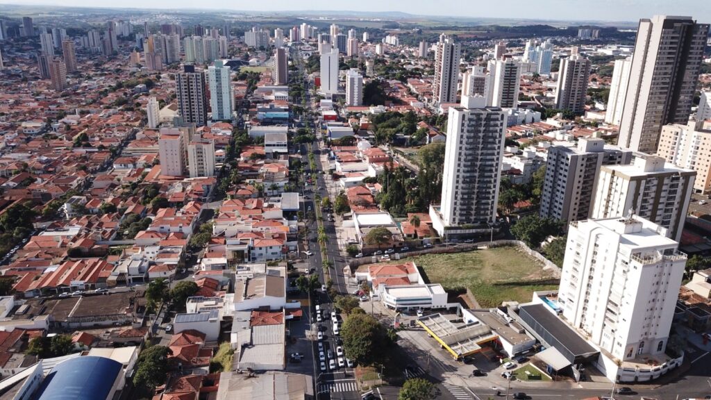A imagem mostra uma vista aérea de uma cidade densa e urbanizada. No centro, uma longa avenida reta corta a paisagem, ladeada por árvores e veículos em movimento. De ambos os lados da avenida, há uma mistura de edifícios residenciais e comerciais. Muitas construções são de baixa altura, com telhados vermelhos característicos, enquanto outras são edifícios altos e modernos, com fachadas de vidro e varandas. A vegetação aparece principalmente ao longo da avenida e em algumas áreas entre os prédios. O céu está claro, indicando um dia ensolarado, e a luz do sol cria sombras definidas, realçando a textura dos telhados e das estruturas. Ao fundo, é possível ver colinas e uma linha de horizonte com mais edifícios, sugerindo que a cidade está situada em uma região levemente ondulada. A imagem transmite a sensação de uma cidade grande, moderna e bem estruturada, com uma combinação de construções antigas e novas.