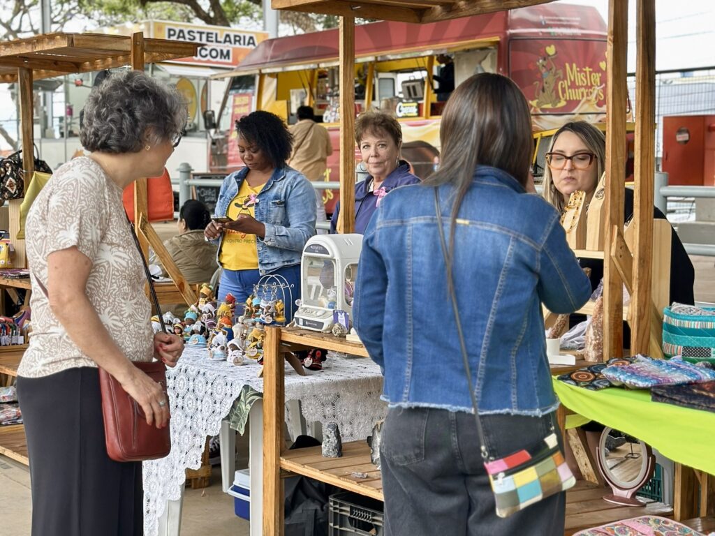 A imagem retrata uma cena em uma feira ao ar livre, possivelmente um mercado de artesanato ou de produtos locais. Várias pessoas estão presentes, interagindo com as mercadorias expostas em bancas de madeira. No primeiro plano, à esquerda, uma senhora de cabelos grisalhos e óculos observa uma banca. Ela veste uma blusa estampada e carrega uma bolsa marrom a tiracolo. Ao seu lado, uma mulher negra, com uma blusa amarela e jaqueta jeans, está concentrada em seu celular. A banca à frente delas exibe uma variedade de pequenos objetos decorativos, possivelmente lembrancinhas ou miniaturas, dispostos sobre uma toalha de crochê branca. Há também uma vitrine transparente, possivelmente para exposição de itens mais delicados. Ao fundo, um food truck com a inscrição "PASTELARIA .COM" sugere a venda de alimentos. Outras pessoas circulam pela feira, e mais bancas com mercadorias diversas, como tecidos coloridos e objetos de decoração, podem ser vistas. A iluminação sugere um dia claro, com luz natural.