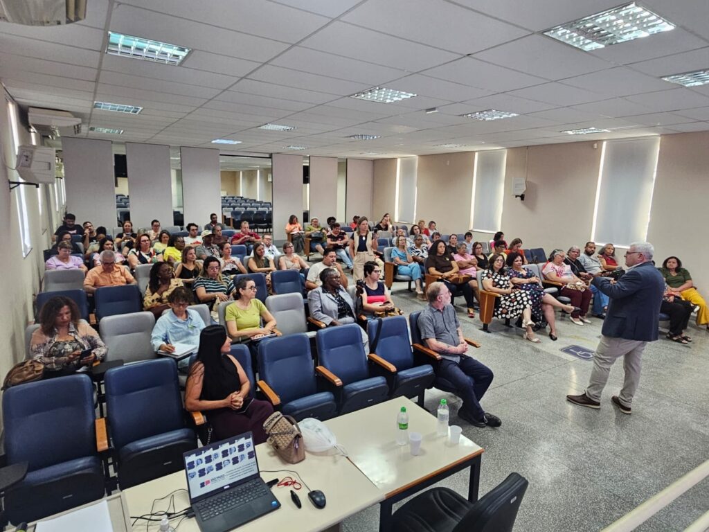 A imagem mostra uma sala de aula ou auditório com várias pessoas sentadas em cadeiras, assistindo a uma apresentação. Há um palestrante em pé na frente da sala, de costas para a câmera, gesticulando com as mãos enquanto fala. As pessoas na plateia parecem atentas, com algumas delas anotando em cadernos. Há um laptop aberto em uma mesa na frente, exibindo uma apresentação com fotos e texto. A sala é iluminada por luzes fluorescentes no teto e há janelas com persianas nas paredes. A atmosfera parece ser de um evento educacional ou profissional.