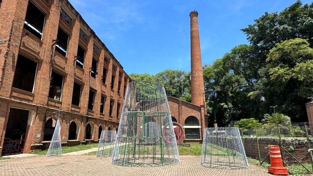 A imagem mostra a fachada de um edifício de tijolos com janelas quebradas e sem vidros. Na frente do edifício, há uma chaminé de tijolos alta e árvores verdes. No chão, há várias estruturas cônicas feitas de metal e com luzes, que parecem ser decorações de Natal. Há também um cone de trânsito laranja e branco em primeiro plano. O céu está azul e limpo, sugerindo um dia ensolarado.