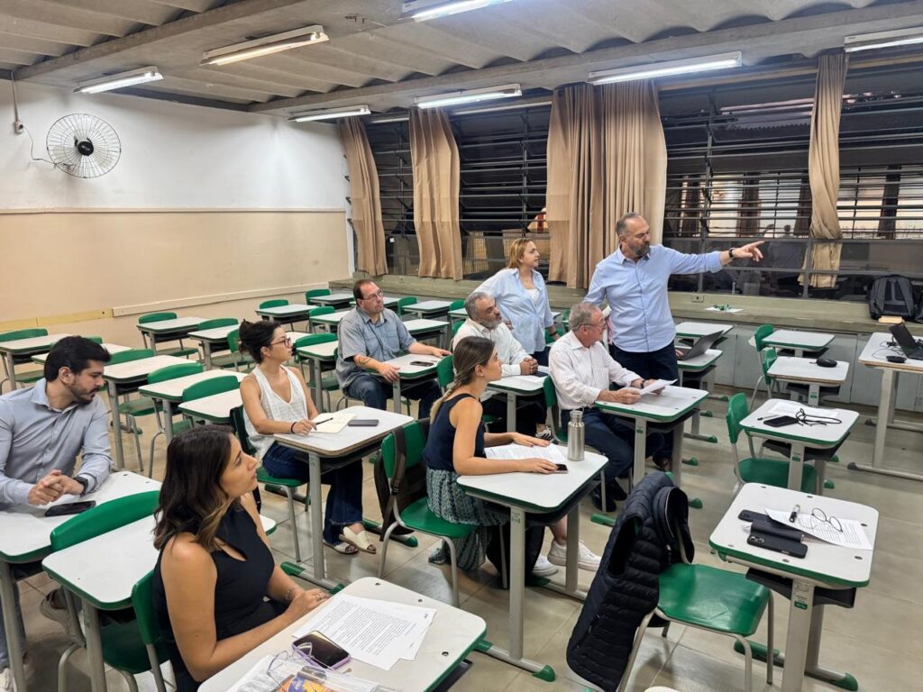 A imagem mostra uma sala de aula com alunos sentados em carteiras e um homem em pé, gesticulando para a frente, como se estivesse a dar uma aula ou apresentação. As carteiras são verdes e brancas, e estão dispostas em filas. A maioria dos alunos está a olhar para o homem em pé, e alguns deles parecem estar a tomar notas. O homem em pé está a usar uma camisa azul e calças escuras. Ele parece estar a falar com os alunos e a apontar para algo à frente deles. A sala de aula tem janelas grandes com cortinas, e há um ventilador de teto. A iluminação é feita por lâmpadas fluorescentes no teto. A atmosfera geral da imagem sugere um ambiente de aprendizagem ou de reunião académica.