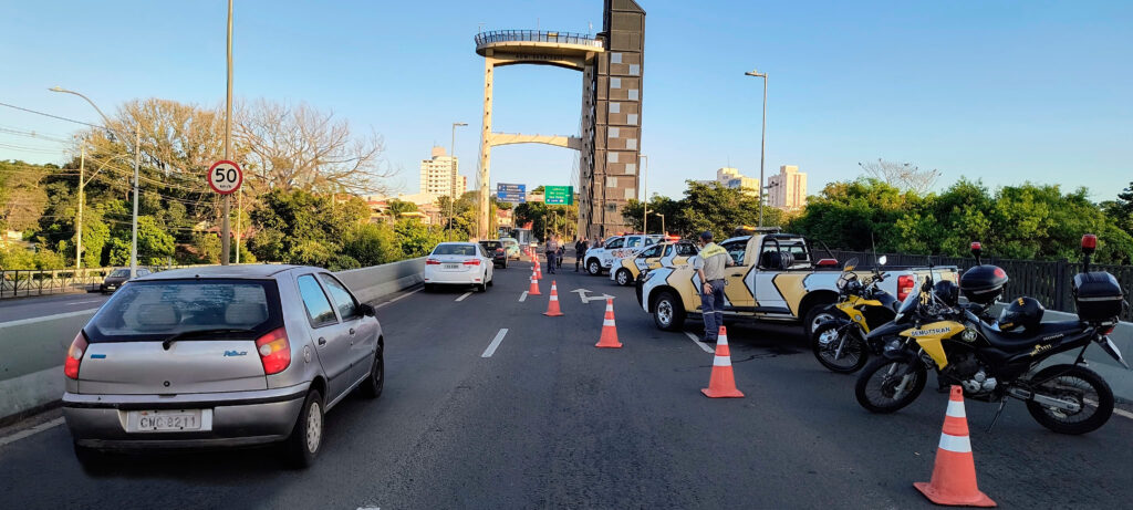 Na imagem, observa-se uma operação de fiscalização de trânsito realizada sobre uma ponte ou via elevada. Cones laranja delimitam a área, conduzindo os veículos para uma faixa de inspeção onde agentes interagem com motoristas. Entre os veículos presentes, destacam-se um Fiat Palio prateado, caminhonetes oficiais brancas e amarelas e duas motocicletas amarelas identificadas com “Trânsito”. Um agente conversa com um condutor, enquanto ao fundo aparecem placas de sinalização, incluindo o limite de velocidade de 50 km/h, além de uma estrutura alta com plataforma de observação. O cenário, em plena luz do dia e cercado por árvores e prédios urbanos, transmite a ideia de controle e segurança viária.
