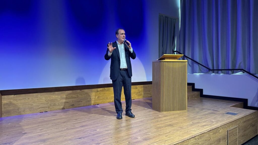 O homem, vestindo um terno escuro, está em um palco de madeira, segurando um microfone em sua mão direita e gesticulando com a mão esquerda. Ele está falando para uma audiência, com um fundo azul vibrante e cortinas azuis escuras à direita. Há um púlpito de madeira com ripas verticais à sua direita, e uma escada desce do lado direito do palco. A iluminação é suave, com tons azuis predominando no fundo e na área atrás do púlpito. O chão do palco é de madeira clara, com um padrão de tábuas de madeira.