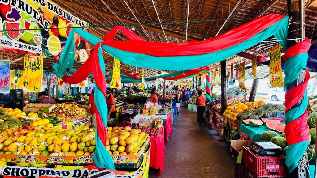 A imagem retrata um vibrante mercado de frutas, com uma abundância de produtos frescos expostos em barracas. O teto é coberto por uma estrutura de telhado de palha, e bandeiras coloridas de tecido vermelho e verde-água estão penduradas, criando uma atmosfera festiva. As barracas estão repletas de uma variedade de frutas, incluindo mangas, mamões, abacaxis e melancias, todos dispostos de forma convidativa. Há também placas com preços e nomes das frutas, como "Mamão Papaya", indicando que se trata de um local de comércio ativo. Pessoas circulam pelo mercado, algumas comprando e outras trabalhando, adicionando vida à cena. A iluminação natural e a disposição organizada das frutas criam um ambiente convidativo e apetitoso.