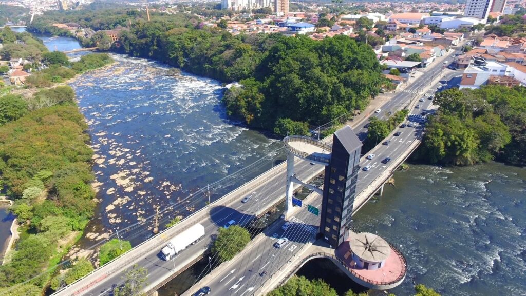 A imagem mostra uma vista aérea de uma ponte sobre um rio caudaloso. A ponte é larga e abriga tráfego de veículos, incluindo um caminhão branco, carros e ônibus. Há uma estrutura arquitetônica moderna e alta na lateral da ponte, com um elevador de vidro e uma plataforma de observação circular. O rio é largo e agitado, com corredeiras e algumas pedras expostas. As margens do rio são cobertas por vegetação exuberante e densa, com árvores altas e folhagem verde. Ao fundo, avista-se uma cidade com edifícios residenciais e comerciais, alguns de vários andares, e telhados de casas. O céu está claro e azul, indicando um dia ensolarado.
