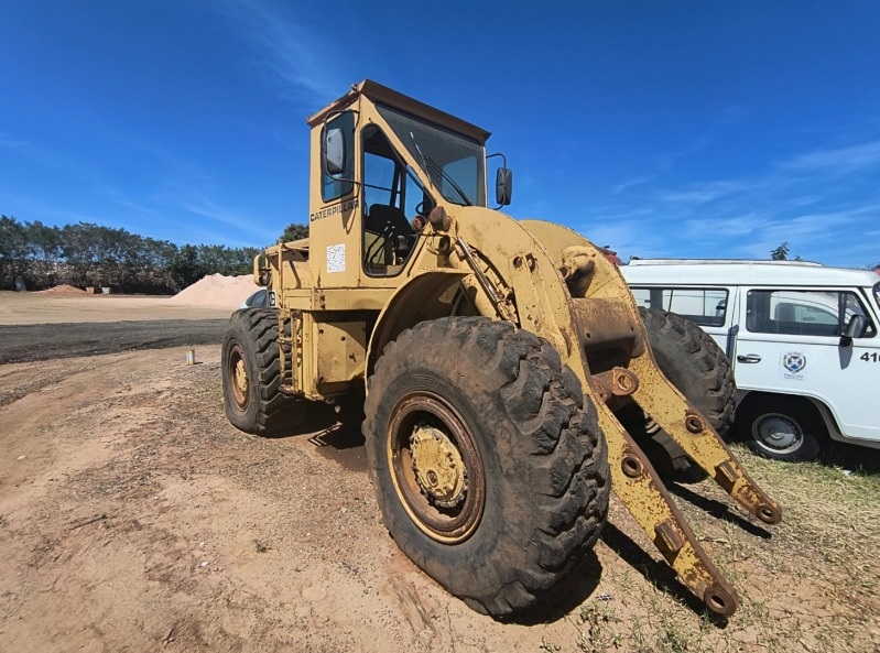 A imagem mostra um veículo de construção pesado da marca Caterpillar, do tipo pá carregadeira, estacionado sobre um terreno de terra. O equipamento possui pneus grandes e robustos, próprios para uso em obras, e apresenta sinais de desgaste, como ferrugem e sujeira. Os braços dianteiros estão estendidos, embora o balde não esteja visível.