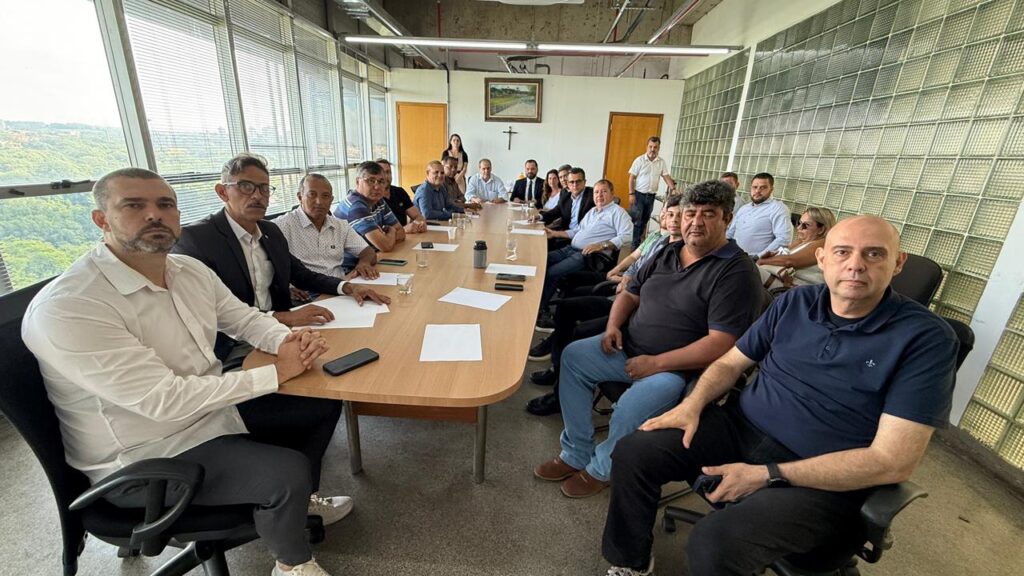 A fotografia mostra uma reunião formal em uma ampla sala de conferências, onde cerca de vinte pessoas estão reunidas em torno de uma grande mesa retangular. Algumas estão sentadas, outras de pé, sugerindo um encontro importante ou decisório. O ambiente é iluminado por janelas que revelam uma paisagem verde ao fundo, enquanto uma parede lateral é composta por blocos de vidro. Na parede oposta, acima de duas portas de madeira, há um crucifixo e um quadro emoldurado, indicando que o espaço pode pertencer a uma instituição religiosa ou ligada a valores tradicionais.