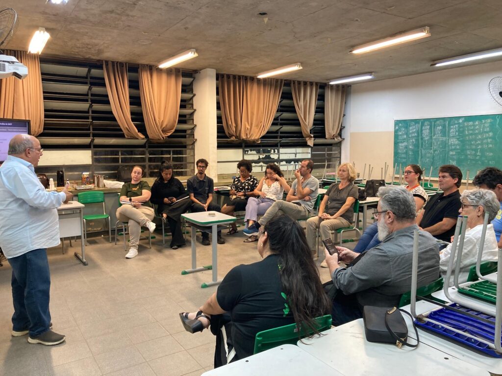 A imagem mostra uma sala de aula com um grupo de pessoas sentadas em cadeiras, a maioria delas voltada para um homem mais velho que está em pé no lado esquerdo da sala, de frente para um projetor. O homem em pé, vestindo uma camisa azul clara e jeans, parece estar dando uma palestra ou apresentação. As pessoas sentadas estão dispostas em um semicírculo, prestando atenção ao palestrante. Há mesas e cadeiras em estilo escolar espalhadas pela sala. Ao fundo, há janelas com cortinas e um quadro negro com algumas anotações escritas. A iluminação da sala vem de luminárias fluorescentes no teto. A atmosfera geral sugere um ambiente de aprendizado ou reunião.