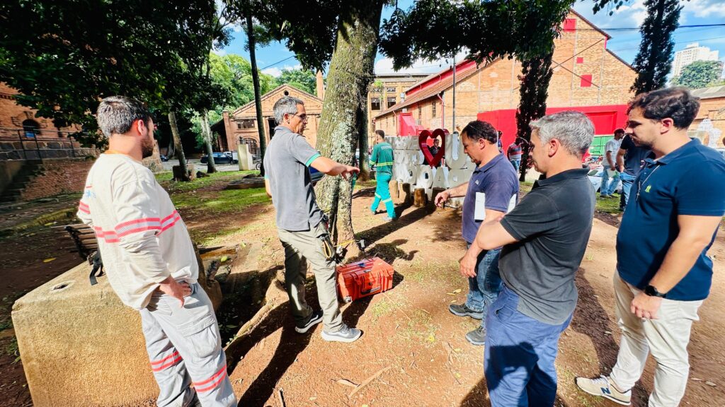 A imagem mostra um grupo de homens em um ambiente externo, possivelmente um parque ou área de lazer, com edifícios de tijolos vermelhos ao fundo. Há árvores e vegetação, com luz solar forte criando sombras no chão. No primeiro plano, um homem com uma camisa de manga comprida bege e calças cinzas, com detalhes em laranja e branco, está virado de costas para a câmera, com as mãos nos quadris. Mais ao centro, um homem com óculos e uma camisa cinza está gesticulando em direção a uma árvore. Ao lado dele, há uma caixa laranja no chão. Outros homens estão em pé, conversando. Ao fundo, um homem com um uniforme verde caminha. Há também uma estrutura grande em forma de coração e letras que parecem formar uma palavra. A arquitetura dos edifícios sugere um local com história, possivelmente uma antiga fábrica ou complexo industrial revitalizado.