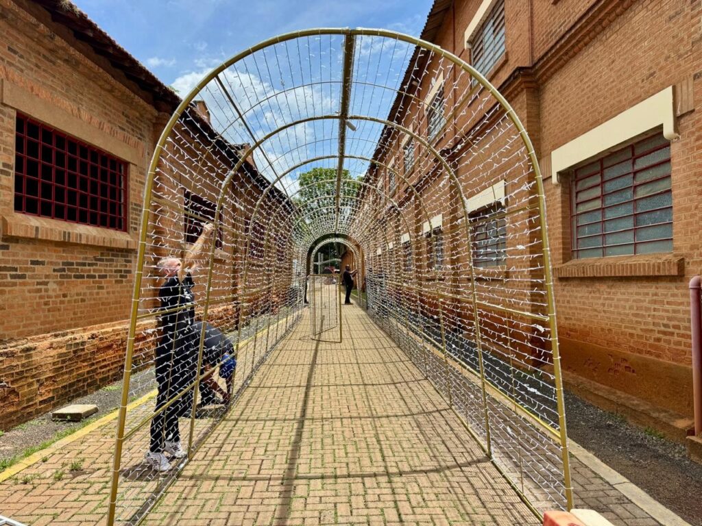 O túnel é feito de uma estrutura metálica amarela e coberto com fios de luzes brancas, que ainda não foram totalmente instaladas. O chão é pavimentado com tijolos e ladeado por edifícios de tijolos vermelhos com janelas de vidro em tons de vermelho. Duas pessoas estão visíveis, trabalhando na instalação das luzes. Uma delas, vestida de preto, está em pé e parece estar ajustando os fios no topo do túnel. A outra pessoa, vestida de azul e preto, está agachada, trabalhando na parte inferior da estrutura. A luz do dia ilumina a cena, criando sombras no chão e realçando a textura dos tijolos dos edifícios. O céu está parcialmente nublado, com algumas nuvens brancas visíveis. A perspectiva da imagem, tirada de dentro do túnel, dá uma sensação de profundidade e convida o espectador a percorrer o caminho iluminado.