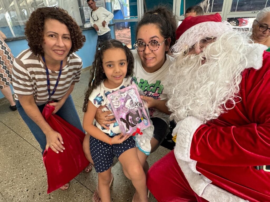 A imagem mostra um grupo de pessoas reunidas, com destaque para uma menina que segura um presente e um homem vestido de Papai Noel. À esquerda, uma mulher sorri para a câmera, segurando uma grande sacola vermelha. Ao lado dela, a menina, com cabelos cacheados, segura um presente embalado em plástico transparente, com detalhes em rosa e azul, e um laço vermelho. Ela veste uma blusa estampada com desenhos e shorts de bolinhas azuis. À direita da menina, uma jovem com óculos redondos e cabelos presos em um rabo de cavalo também sorri. Ela veste uma blusa branca com uma estampa verde que diz "itch friend". O homem vestido de Papai Noel, com sua barba branca característica e gorro vermelho, está posicionado à direita, interagindo com as crianças. Ele veste o traje vermelho com detalhes brancos. Ao fundo, outras pessoas podem ser vistas, incluindo um homem em pé e uma mulher mais velha com óculos. O ambiente parece ser um local público, possivelmente uma instituição ou evento, com paredes azuis e piso claro. A atmosfera geral da imagem é de alegria e celebração.