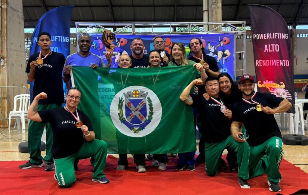 A imagem mostra um grupo de pessoas celebrando uma conquista em um evento de powerlifting. No centro da imagem, há uma bandeira verde com o brasão da cidade de Piracicaba. As pessoas, vestindo camisetas pretas com a inscrição "PIRACICABA" e calças verdes, posam para a foto segurando a bandeira e exibindo medalhas. Algumas delas fazem poses de força, demonstrando orgulho e satisfação. Ao fundo, há banners com o logo "POWERLIFTING ALTO RENDIMENTO" e ilustrações relacionadas ao esporte. A atmosfera geral é de celebração e sucesso, com os participantes claramente felizes com seus resultados.