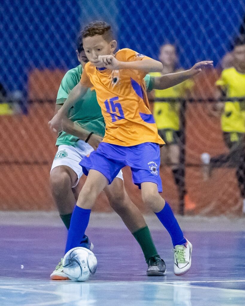 A imagem mostra dois jovens jogadores de futebol de salão em ação durante uma partida. O jogador em primeiro plano, vestindo uma camisa laranja com o número 15 em azul e shorts azuis, está com a bola nos pés, em movimento. Ele tem cabelos castanhos curtos e está focado no jogo. O outro jogador, parcialmente visível atrás dele, veste uma camisa verde e shorts brancos. Ambos os jogadores usam meias de cores diferentes: o jogador da frente usa meias azuis, enquanto o jogador de trás usa meias verdes. Os tênis de futebol de salão de ambos os jogadores são visíveis, com detalhes distintos. O fundo da imagem é desfocado, mostrando uma quadra com uma rede azul e espectadores ao longe, indicando que a partida está acontecendo em um ginásio.