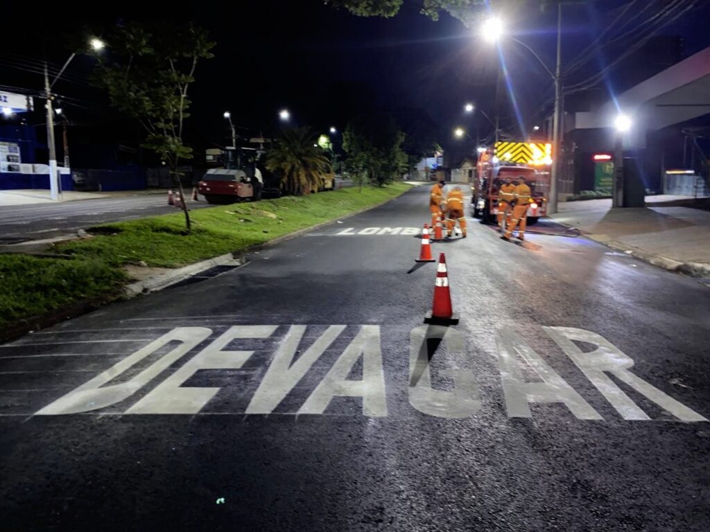 A imagem mostra uma cena noturna em uma rua, onde trabalhadores uniformizados em laranja estão realizando algum tipo de serviço na pista. Há um caminhão com luzes de advertência piscando ao fundo, e cones de trânsito laranja estão espalhados pela via. Na parte inferior da imagem, a palavra "DEVA*GA*R" está pintada em letras brancas grandes e estilizadas diretamente no asfalto recém-aplicado ou limpo. A pintura da palavra ainda parece fresca, com algumas áreas brilhantes, sugerindo que o trabalho de sinalização está em andamento ou acabou de ser concluído. A iluminação pública e as luzes dos veículos criam um ambiente de trabalho noturno. Ao lado da rua, há grama e árvores, e ao fundo, edifícios e postes de luz.
