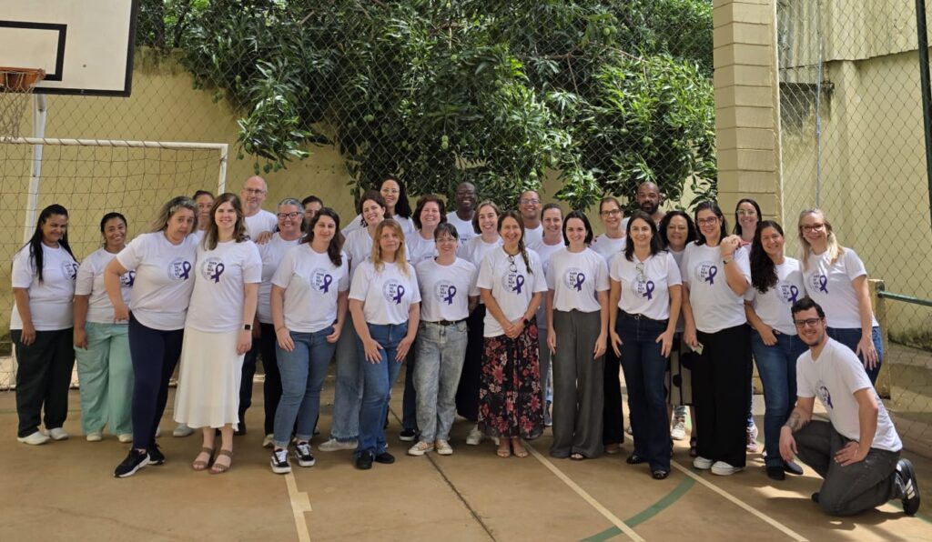 A imagem mostra um grupo de cerca de trinta pessoas reunidas em uma quadra de basquete ao ar livre, todas vestindo camisetas brancas com o símbolo de um laço roxo.