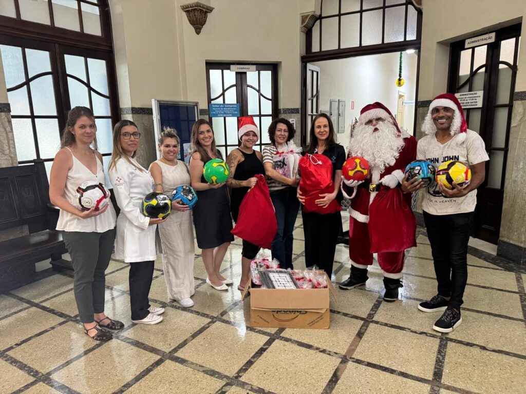 A imagem mostra um grupo de pessoas posando para uma foto em um corredor com um piso de mármore. No centro, há um homem vestido de Papai Noel, segurando sacos vermelhos. Ao lado dele, um homem com um chapéu de Papai Noel e uma camiseta com a estampa "JOHN JOHN". À esquerda do Papai Noel, estão sete mulheres. Uma delas usa um jaleco branco e segura uma bola de futebol preta e amarela. Outra mulher segura uma bola de futebol verde e preta. As outras mulheres seguram sacos vermelhos. No chão, há uma caixa de papelão contendo vários pacotes embrulhados em papel celofane, possivelmente presentes. Há também uma caixa com um tabuleiro de jogo. O ambiente parece ser um hospital ou uma instituição pública, com portas e sinalizações ao fundo.