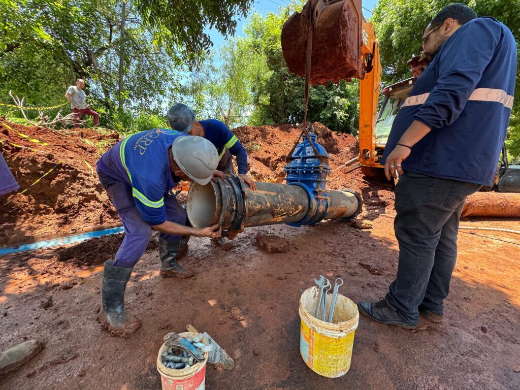 A imagem mostra um cenário de construção ao ar livre, onde três homens estão trabalhando na instalação de uma grande válvula industrial em um cano metálico grosso. O chão é de terra vermelha, e há grandes montes dessa terra ao redor, indicando que o local foi escavado recentemente. No centro da imagem, um grande cano enferrujado está apoiado, enquanto uma válvula azul vibrante está sendo posicionada sobre ele, segurada por uma escavadeira laranja, cuja pá está levantada para sustentar a válvula. Dois dos trabalhadores estão próximos ao cano, vestindo uniformes azuis com faixas refletivas nas mangas, capacetes de segurança e botas. Um deles está agachado, aparentemente ajustando ou conectando o cano, enquanto o outro observa ou ajuda. Um terceiro homem, vestido com uma camisa azul de manga longa e calça escura, está de pé ao lado, olhando para a válvula, usando pulseiras e anéis. Ao fundo, há árvores e vegetação verde, contrastando com a terra vermelha do solo. Também é possível ver uma fita amarela de segurança e um balde amarelo com algumas ferramentas. A escavadeira tem a marca "CASE" visível na lateral. A cena é iluminada por luz solar forte, que destaca as cores vibrantes e cria sombras marcantes, evidenciando a textura da terra e dos equipamentos. O ambiente sugere um projeto de infraestrutura, possivelmente relacionado ao sistema de água ou esgoto, em uma área externa, com bastante trabalho manual e uso de maquinário pesado.