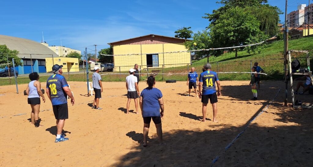 A partida de vôlei de praia está em andamento sob um céu azul claro. Um grupo de pessoas, a maioria homens mais velhos, está jogando em uma quadra de areia. Há uma rede esticada no meio da quadra, e os jogadores estão posicionados em ambos os lados, prontos para o jogo. Alguns jogadores vestem camisetas de time com números e nomes, sugerindo uma partida amigável ou um torneio local. Ao fundo, um prédio amarelo com um telhado marrom se destaca, e árvores verdes e um gramado inclinado criam um cenário natural. A luz do sol forte projeta sombras nítidas na areia, indicando que é um dia ensolarado.