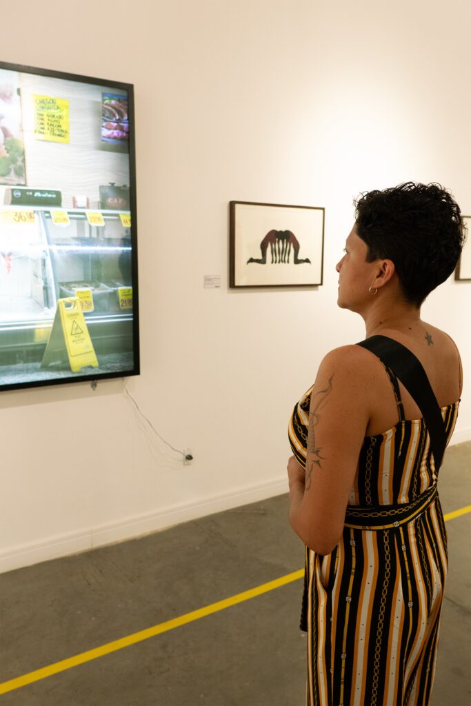 A pessoa na imagem está em uma galeria de arte, admirando uma obra de arte exibida em uma tela. A obra de arte parece ser uma representação de um supermercado ou açougue, com produtos em exposição e preços visíveis. Há também uma segunda obra de arte na parede, uma gravura emoldurada que retrata figuras humanas em uma pose curvada. A pessoa está usando um macacão listrado em amarelo, preto e branco, com uma alça preta sobre o ombro. O chão da galeria é de concreto com uma linha amarela pintada, e a iluminação é suave, criando uma atmosfera contemplativa.