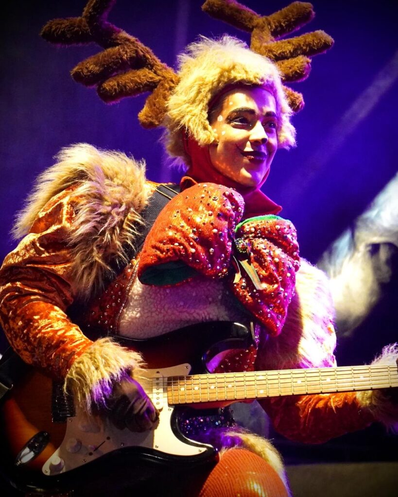 A imagem mostra um músico fantasiado de rena tocando guitarra elétrica em um palco com iluminação roxa. O traje da rena inclui chifres marrons peludos, uma peruca loira fofa, um casaco laranja brilhante com detalhes de pele e luvas vermelhas adornadas com lantejoulas. O músico tem maquiagem facial branca com pontos pretos e um sorriso. A guitarra é uma Fender Stratocaster sunburst. A iluminação cria um ambiente festivo e teatral.