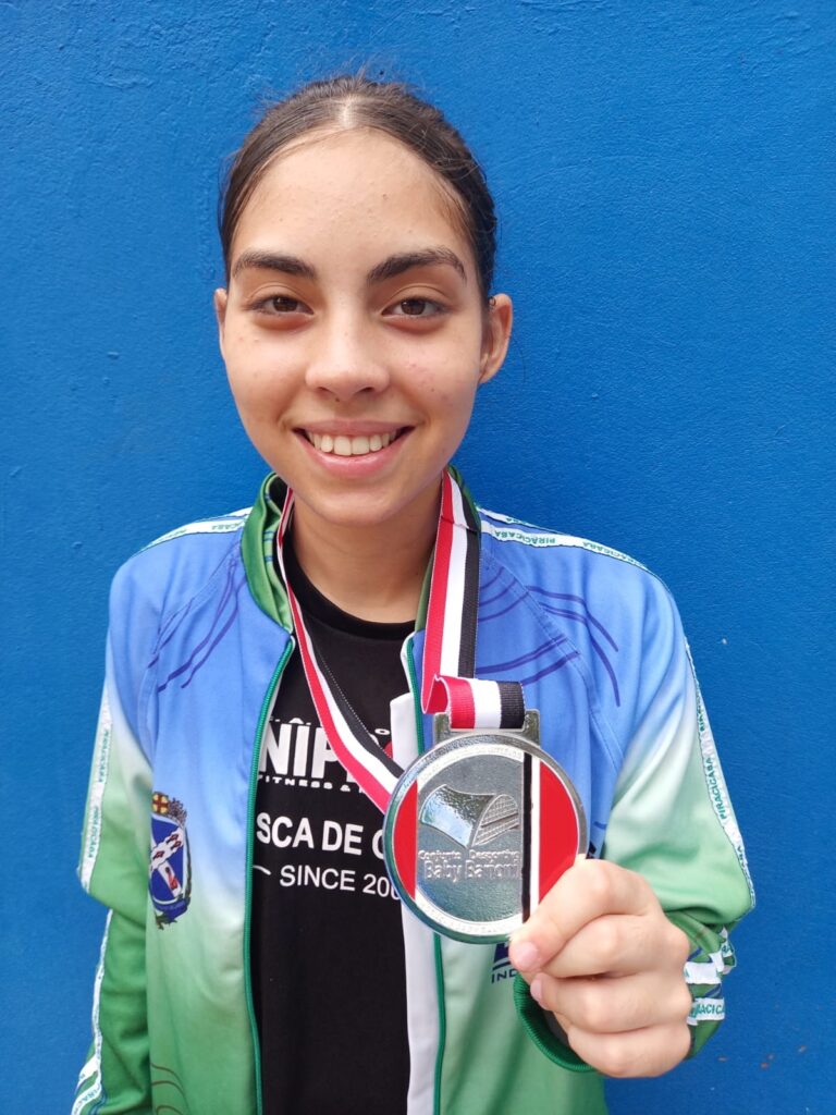 A jovem sorri para a câmera, segurando uma medalha prateada com uma fita tricolor nas cores preto, branco, vermelho e verde. Ela veste uma jaqueta esportiva azul e verde com detalhes em branco e um emblema em seu ombro esquerdo. Por baixo da jaqueta, ela usa uma camiseta preta com texto branco que parece ser um logotipo de academia ou time. O fundo é uma parede azul vibrante. A medalha tem um design circular com uma imagem gravada no centro e o texto "Baby Baroni" visível. O texto na jaqueta e na camiseta indica que ela faz parte de uma organização ou evento relacionado a Piracicaba, possivelmente uma academia ou competição esportiva.