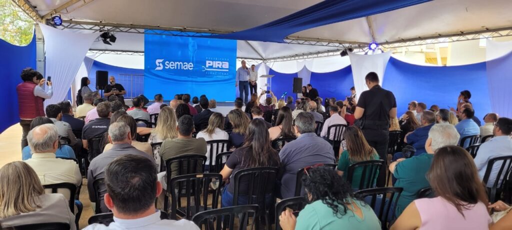 A imagem mostra um evento ao ar livre, possivelmente uma cerimônia ou apresentação, sob uma tenda decorada com tecidos brancos e azuis. Há um palco com um telão ao fundo que exibe o logo "SEMAE PIRA" e o slogan "PREFEITURA PIRA". Duas pessoas estão em pé no palco, uma delas falando. Uma multidão de pessoas está sentada em cadeiras, voltada para o palco, assistindo à apresentação. Algumas pessoas na plateia estão tirando fotos ou gravando com seus celulares. Há também algumas pessoas em pé nas laterais, observando o evento. A atmosfera parece ser formal, mas também descontraída, com a luz do dia iluminando a cena.