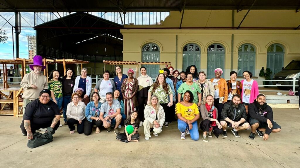 A imagem mostra um grupo grande de pessoas, a maioria mulheres, posando para uma foto em frente a um edifício com janelas azuis e um telhado de metal. O grupo está em um espaço aberto, possivelmente um pátio ou mercado coberto, com estruturas de madeira ao fundo. Muitas das mulheres estão usando fitas cor-de-rosa, um símbolo comum de conscientização sobre o câncer de mama. Algumas pessoas usam camisetas com estampas, e uma pessoa à esquerda usa um chapéu roxo grande e chamativo. O clima geral da foto parece ser de união e celebração. O grupo está posicionado em várias fileiras, com algumas pessoas em pé e outras agachadas ou sentadas no chão. A iluminação sugere que a foto foi tirada durante o dia. Há uma van estacionada ao fundo, à direita.