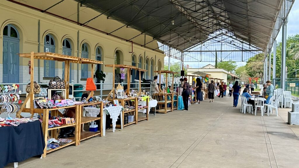 A imagem retrata um mercado ao ar livre, possivelmente uma feira de artesanato ou de antiguidades, sob uma grande cobertura metálica. Diversas barracas de madeira com prateleiras exibem uma variedade de produtos, como bolsas, acessórios de cabelo, carteiras e outros itens artesanais. Pessoas circulam entre as barracas, observando e comprando os produtos. Ao fundo, um edifício com janelas arqueadas e portas azuis serve de cenário, enquanto áreas verdes e árvores complementam a paisagem externa. Cadeiras plásticas brancas empilhadas sugerem um espaço para descanso ou alimentação.