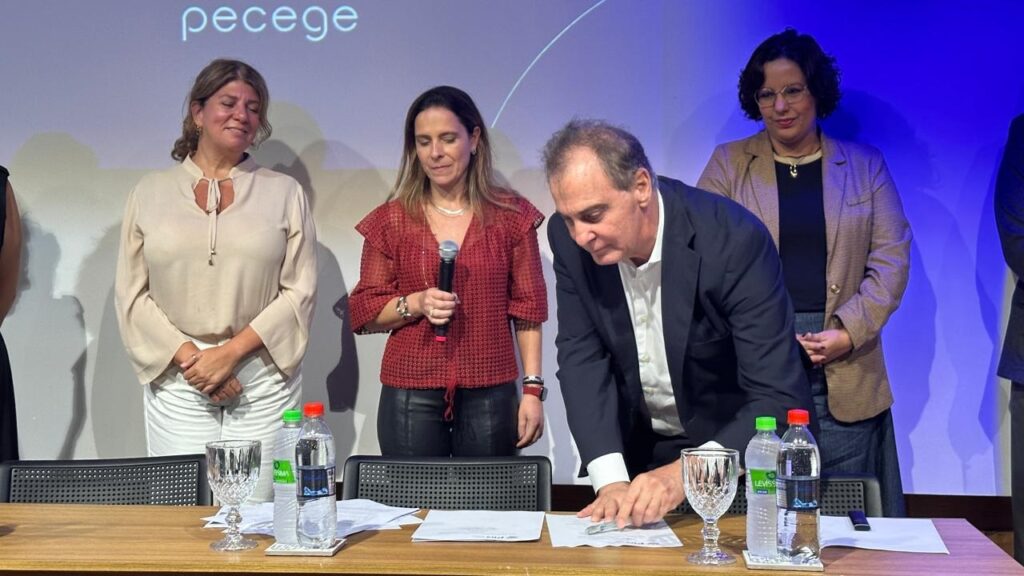 A imagem mostra um grupo de quatro pessoas em pé atrás de uma mesa de madeira, onde há garrafas de água, um copo de cristal e papéis. Ao fundo, um telão exibe a palavra "pecege" em letras brancas.À esquerda, uma mulher loira com um coque baixo veste uma blusa bege clara de mangas compridas com um detalhe de amarração no decote e calças brancas. Ela sorri levemente, com as mãos cruzadas na frente do corpo.Ao lado dela, uma mulher de cabelos castanhos lisos, vestindo uma blusa vermelha com mangas bufantes e calças pretas, segura um microfone preto na mão direita. Ela olha para baixo, em direção à mesa.No centro da imagem, um homem de cabelos grisalhos veste um terno escuro sobre uma camisa branca. Ele está debruçado sobre a mesa, assinando um documento com uma caneta prateada.À direita, uma mulher com óculos e cabelos escuros e cacheados veste um blazer marrom sobre uma blusa preta. Ela olha para frente, com as mãos juntas na frente do corpo.A iluminação é azulada no fundo, proveniente do telão, e mais clara na frente, iluminando o grupo e a mesa. A atmosfera parece ser de um evento formal, possivelmente uma assinatura de acordo ou um anúncio.