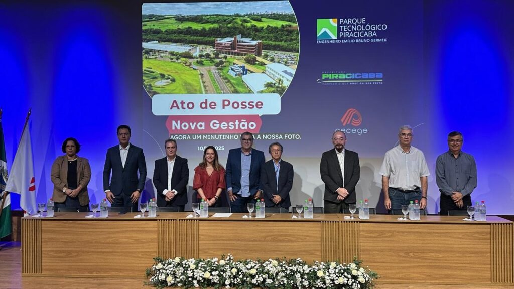 A imagem mostra uma cerimônia de posse, com um grupo de pessoas em pé atrás de uma mesa de madeira, e um telão ao fundo. O telão exibe uma imagem aérea de um complexo de edifícios e áreas verdes, com o logo do "PARQUE TECNOLÓGICO PIRACICABA" e o nome "ENGENHEIRO EMÍLIO BRUNO GERMEK". Abaixo, em destaque, lê-se "Ato de Posse" e "Nova Gestão", com a frase "AGORA UM MINUTINHO PARA A NOSSA FOTO." e a data "10/025". Há também o logo da "PREFEITURA PIRACICABA" e da "recege". As pessoas presentes parecem ser autoridades ou convidados importantes, vestidas formalmente. Há bandeiras à esquerda e arranjos florais na frente da mesa.