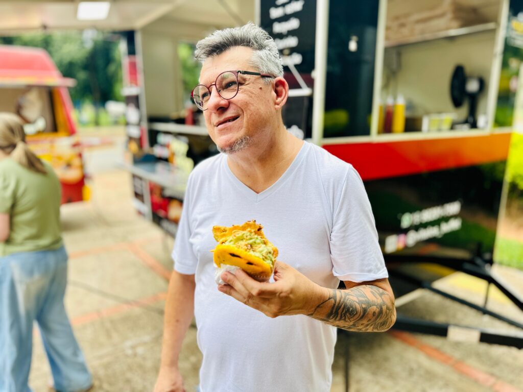 Um homem de meia-idade com cabelo grisalho e óculos de aro vermelho está segurando um pedaço de comida, possivelmente um taco ou arepa, em sua mão direita. Ele está vestindo uma camiseta branca e tem uma tatuagem em seu braço esquerdo. Ele sorri e olha para o lado, como se estivesse conversando com alguém. Ao fundo, há um food truck com uma pintura colorida em tons de vermelho, laranja e amarelo. Outro food truck, vermelho, está visível à esquerda, e uma pessoa com uma camiseta verde e calça jeans está de costas, olhando para o food truck à esquerda. O ambiente parece ser ao ar livre, possivelmente um parque ou uma área de eventos, com árvores e árvores ao fundo. O chão é de concreto com algumas marcações.