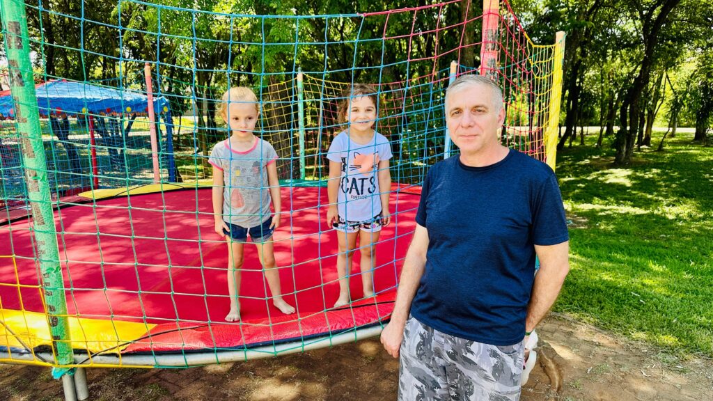 A imagem mostra um homem adulto em primeiro plano, à direita, olhando para a câmera com uma expressão neutra. Ele veste uma camiseta azul escura e calças com estampa camuflada em tons de cinza e branco. Atrás dele, dentro de uma grande cama elástica com rede de proteção colorida (verde, azul, amarela e vermelha), estão duas meninas. A menina da esquerda, com cabelos loiros presos, veste uma camiseta cinza e shorts jeans. A menina da direita, com cabelos castanhos mais curtos, veste uma camiseta branca com estampa de gatos e a palavra "CATS FOREVER", e shorts estampados. Ambas estão descalças. A cama elástica tem uma superfície vermelha com detalhes amarelos nas bordas. A rede de proteção é feita de malha fina e colorida, com postes verticais. Ao fundo, à esquerda, vislumbra-se uma estrutura com um toldo azul e vermelho, possivelmente parte de outra área de lazer. O restante do cenário é um parque com gramado verde e árvores frondosas, sob um céu claro e ensolarado, indicando um dia agradável ao ar livre. A luz do sol cria sombras no gramado, realçando a atmosfera natural do local.
