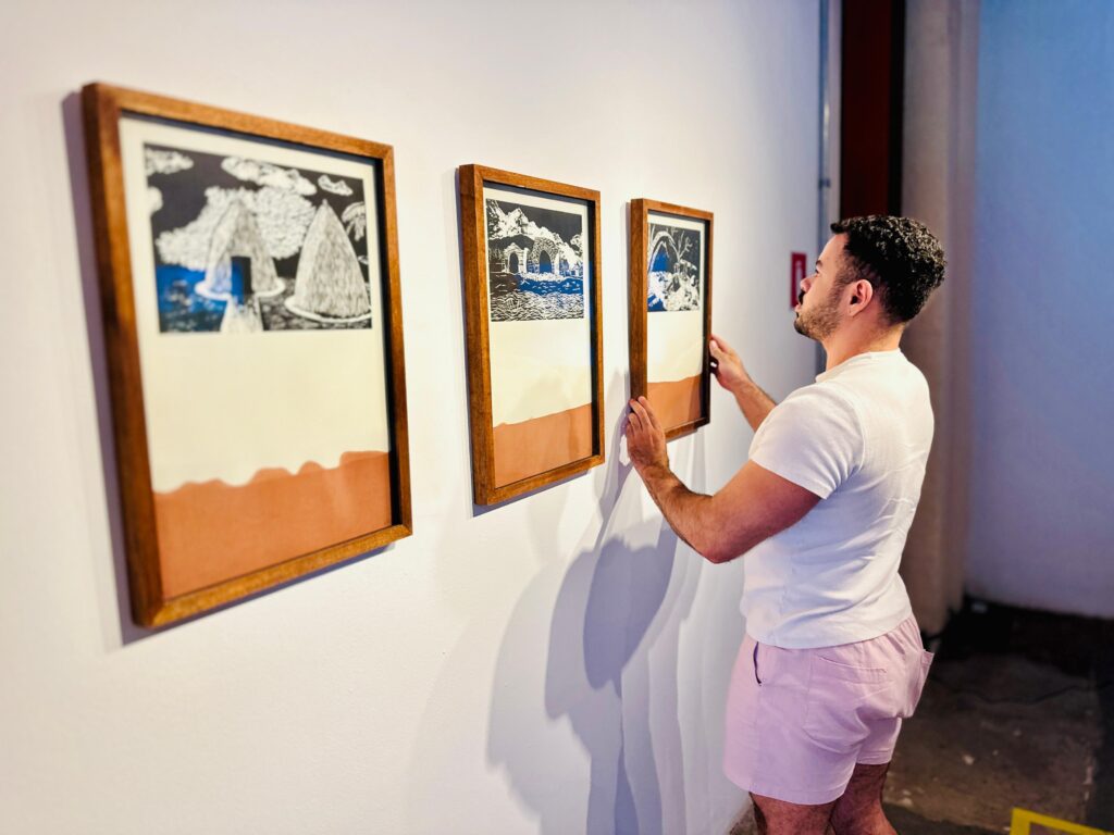 A imagem mostra um homem com barba e cabelo encaracolado, vestindo uma camiseta branca e shorts rosa claro, pendurando um quadro em uma parede branca. Ele está de perfil, olhando para o quadro que está ajustando. Há outros dois quadros pendurados na parede à esquerda dele, todos com molduras de madeira escura e com um estilo de arte semelhante, apresentando paisagens estilizadas em preto e branco e tons terrosos. A iluminação cria sombras projetadas na parede, adicionando profundidade à cena.