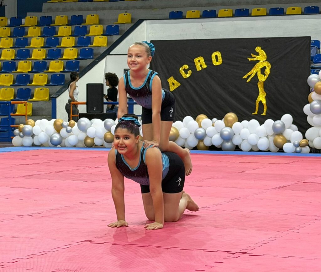 A imagem mostra duas jovens ginastas em uma apresentação. A ginasta de baixo está em posição de quatro apoios em um tapete rosa, enquanto a ginasta de cima está equilibrada em suas costas. Ambas usam trajes de ginástica pretos e azuis. Ao fundo, há arquibancadas com assentos azuis e amarelos. Um banner preto com letras douradas brilhantes que dizem "ACRO" está pendurado atrás das ginastas, com uma silhueta dourada de duas pessoas em uma pose acrobática. Uma fileira de balões brancos, dourados e prateados decora a base do palco. Várias pessoas podem ser vistas nas arquibancadas e ao fundo, incluindo uma pessoa perto de um alto-falante.