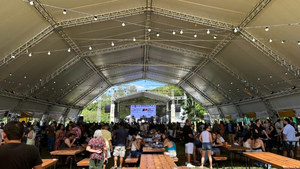 A imagem mostra um grande evento ao ar livre, possivelmente um festival ou feira, sob uma tenda expansiva com estrutura metálica aparente. A tenda é iluminada por uma série de lâmpadas penduradas, criando uma atmosfera festiva. No fundo, um palco com um telão exibe imagens e há uma banda se apresentando. A área sob a tenda está repleta de pessoas, a maioria em pé ou sentada em mesas de madeira compridas, sugerindo um ambiente de convivência e consumo de alimentos e bebidas. A vegetação verde e árvores visíveis ao fundo, juntamente com o céu azul, indicam que o evento ocorre durante o dia em um local aberto. Há também banners e sinalizações nas laterais da tenda, típicos de eventos comerciais ou culturais.