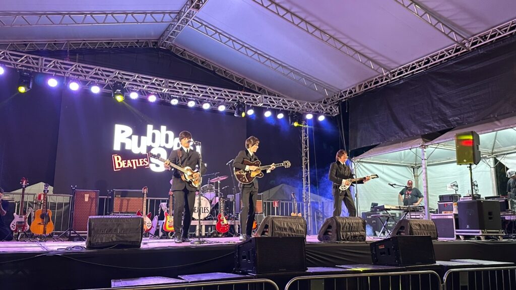 Uma banda tributo aos Beatles, chamada "Rubber Soul Beatles", se apresenta em um palco. Os quatro membros da banda estão vestidos com ternos pretos e perucas que imitam os penteados dos Beatles. Eles estão tocando instrumentos como guitarras e bateria. Atrás deles, há um telão com o nome da banda em destaque. A iluminação do palco é vibrante, com luzes roxas e amarelas, criando uma atmosfera de show. Há também equipamentos de som, amplificadores e outros instrumentos espalhados pelo palco. Ao fundo, algumas pessoas da equipe técnica podem ser vistas.