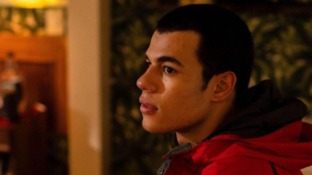 A imagem mostra um close-up do perfil de um jovem com pele morena e cabelo curto e escuro. Ele está vestindo uma jaqueta vermelha com detalhes em cinza escuro. A luz incide sobre o lado direito do seu rosto, destacando seus olhos escuros, nariz proeminente e lábios cheios. O fundo está desfocado, com tons quentes de laranja e verde, sugerindo um ambiente interno com iluminação ambiente. A expressão do jovem parece pensativa ou atenta a algo fora do quadro.