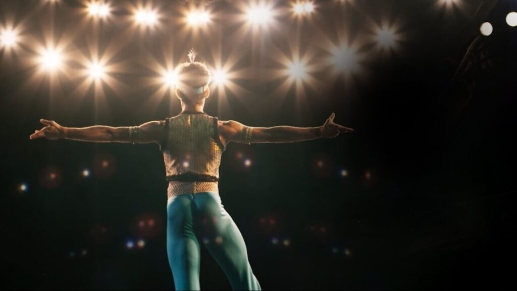 A imagem mostra um artista de circo em um palco, de costas para a câmera. Ele está usando um traje cintilante e azul-petróleo, com braços abertos e um pé ligeiramente levantado, como se estivesse no meio de uma performance. O palco é iluminado por uma série de holofotes brilhantes que criam um efeito de estrela ao redor das luzes. O fundo é escuro, contrastando com a iluminação vibrante do palco e o traje do artista. Há também alguns pontos de luz difusos no fundo, que podem ser outros refletores ou efeitos especiais. A pose do artista sugere movimento e expressão artística, capturando um momento dinâmico de sua apresentação.