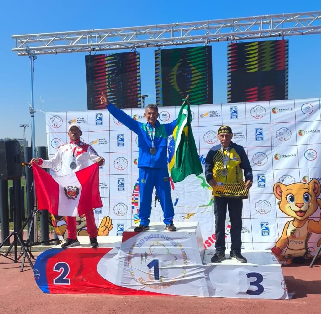 A imagem mostra três homens em um pódio de premiação, celebrando suas vitórias em uma competição de atletismo. O pódio é feito de metal branco e tem os números "1", "2" e "3" em destaque. O homem no centro, no degrau mais alto (posição 1), é um atleta brasileiro, vestindo um uniforme azul com detalhes verdes e amarelos, segurando a bandeira do Brasil. Ele tem uma medalha de prata no pescoço e está com o braço levantado em sinal de vitória. À sua esquerda, no degrau inferior (posição 2), está um atleta peruano, vestindo um uniforme branco e vermelho, segurando a bandeira do Peru. Ele também ostenta uma medalha de prata e parece estar posando para a foto. À direita do atleta brasileiro, no degrau inferior (posição 3), está um atleta colombiano, vestindo um uniforme amarelo e preto. Ele segura uma placa com o nome "COLOMBIA" e também exibe uma medalha de prata.