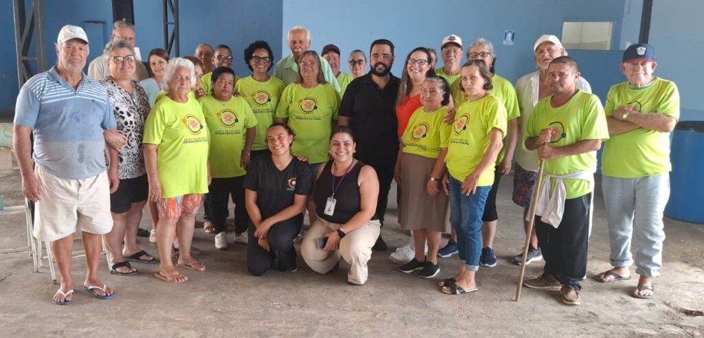 A imagem mostra um grupo de pessoas, a maioria idosas, reunidas em um ambiente que parece ser um espaço comunitário ou de lazer. Várias pessoas no centro da imagem vestem camisetas verdes com um logotipo circular que parece ser de uma organização ou evento. Há também homens e mulheres mais novos no grupo, incluindo um homem com barba que está de pé atrás de algumas mulheres mais velhas, e duas mulheres ajoelhadas na frente. O homem mais à esquerda, vestindo uma camisa polo azul e branca e shorts brancos, parece ser o mais proeminente na frente. Ao lado dele, uma mulher com óculos e uma blusa estampada. Mais atrás, um homem com uma camisa clara e um boné branco, e à sua direita, um homem com uma camisa verde e um boné azul, com os braços cruzados. As pessoas estão em diferentes posições, algumas em pé e outras ajoelhadas, e a atmosfera geral parece ser amigável e casual. O fundo é uma parede azul clara e uma estrutura metálica.