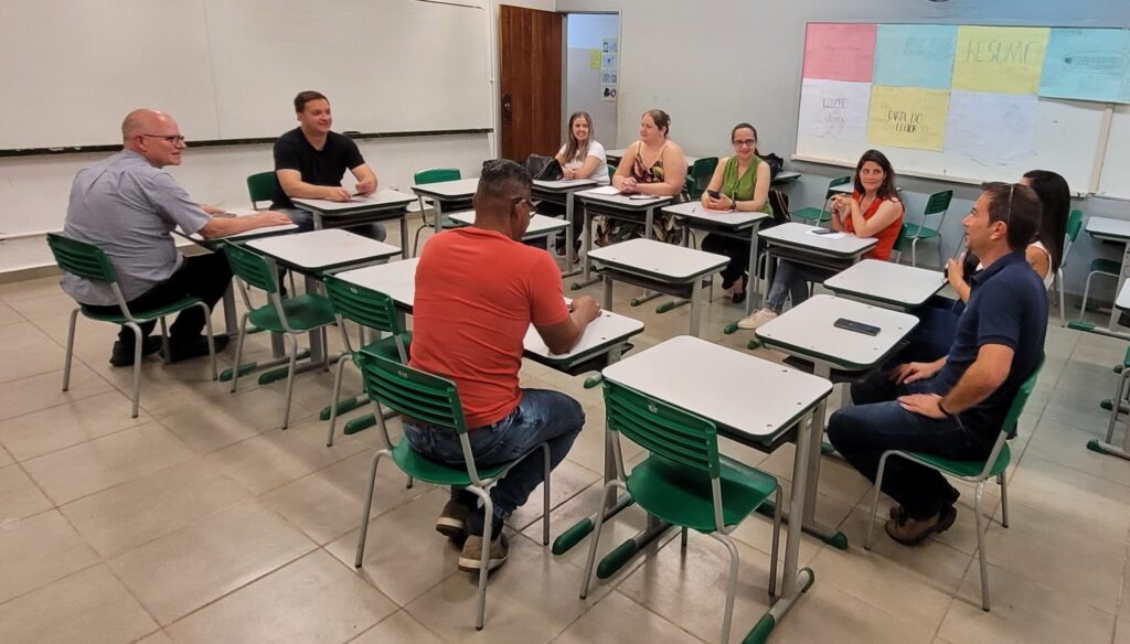 A imagem mostra um grupo de pessoas sentadas em carteiras em sala de aula. As carteiras são duplas e as cadeiras são verdes. Ao fundo, há um quadro branco com anotações e desenhos. As pessoas parecem estar participando de uma aula ou reunião. Há sete pessoas visíveis na imagem, todas sentadas em carteiras. As pessoas estão vestidas casualmente, com algumas usando camisas e outras camisetas. A sala de aula parece ser pequena e tem um piso de ladrilho. Há uma porta no lado direito da sala e uma janela no lado esquerdo. O quadro branco ao fundo está coberto de papéis coloridos com anotações. A iluminação na sala é suave e uniforme, vindo de uma fonte de luz acima. As sombras projetadas pelas carteiras e pessoas sugerem que a luz está vindo de cima e ligeiramente para a direita. A imagem parece ter sido tirada de um ângulo ligeiramente elevado, olhando para baixo nas pessoas e carteiras.