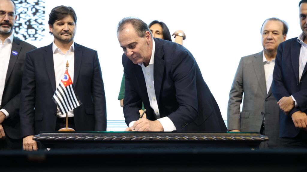 A imagem mostra um grupo de homens em um evento oficial, possivelmente uma cerimônia de assinatura de algum documento. O foco principal está em um homem no centro, vestindo um terno azul escuro e camisa branca, que está abaixado assinando um documento sobre uma mesa ornamentada. Ao lado dele, à esquerda, há um pequeno mastro com a bandeira do estado de São Paulo. Outros homens, também vestidos formalmente com ternos, estão em pé ao redor, observando a assinatura. O fundo é claro, com uma projeção abstrata em tons de branco e preto à esquerda. A iluminação sugere um evento em ambiente interno.