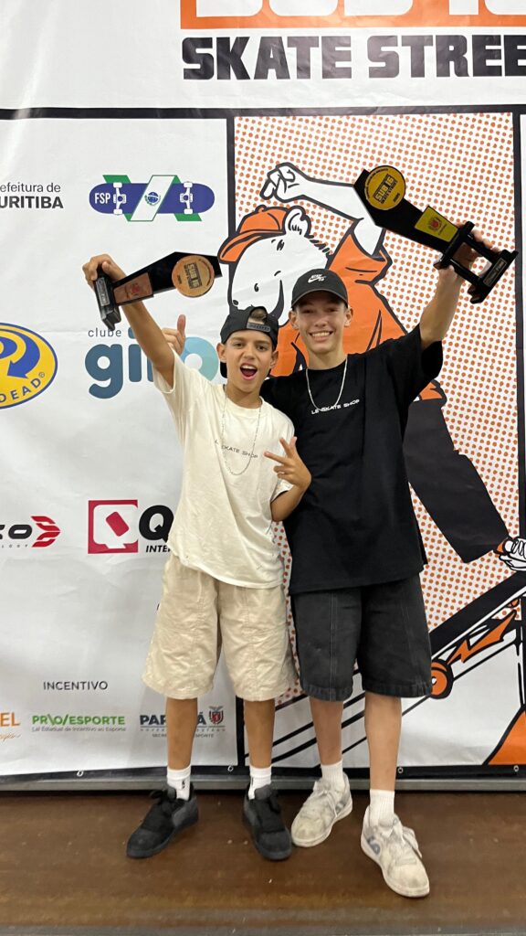 A imagem mostra dois jovens skatistas sorrindo e segurando troféus em um evento de skate. O skatista à esquerda usa uma camiseta branca e shorts bege, enquanto o da direita veste uma camiseta preta e shorts escuros. Ambos usam bonés e colares. Ao fundo, há um banner com o título "SKATE STREET" e logotipos de patrocinadores, incluindo "Prefeitura de Curitiba" e "FSP". Os jovens parecem estar comemorando suas conquistas no evento.