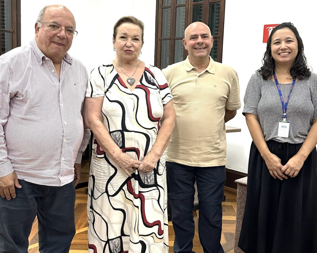 A imagem mostra quatro pessoas em pé, possivelmente em um evento ou reunião. Da esquerda para a direita, vemos um homem mais velho com óculos, vestindo uma camisa listrada em tons de rosa e branco e calças azuis escuras. Ao seu lado, uma mulher de meia-idade com um colar em forma de coração, usando um vestido estampado em branco com linhas pretas e vermelhas. Em seguida, um homem careca com uma camisa polo bege e calças azuis escuras. Por fim, uma mulher mais jovem com cabelos escuros e cacheados, vestindo uma blusa estampada em preto e branco e uma saia preta longa, com um crachá pendurado no pescoço. Todos sorriem para a câmera, transmitindo uma atmosfera amigável e profissional. Ao fundo, observa-se uma janela com madeira escura e uma parede branca, além de mesas e uma placa vermelha no canto superior direito. O piso é de madeira com um padrão em espinha.