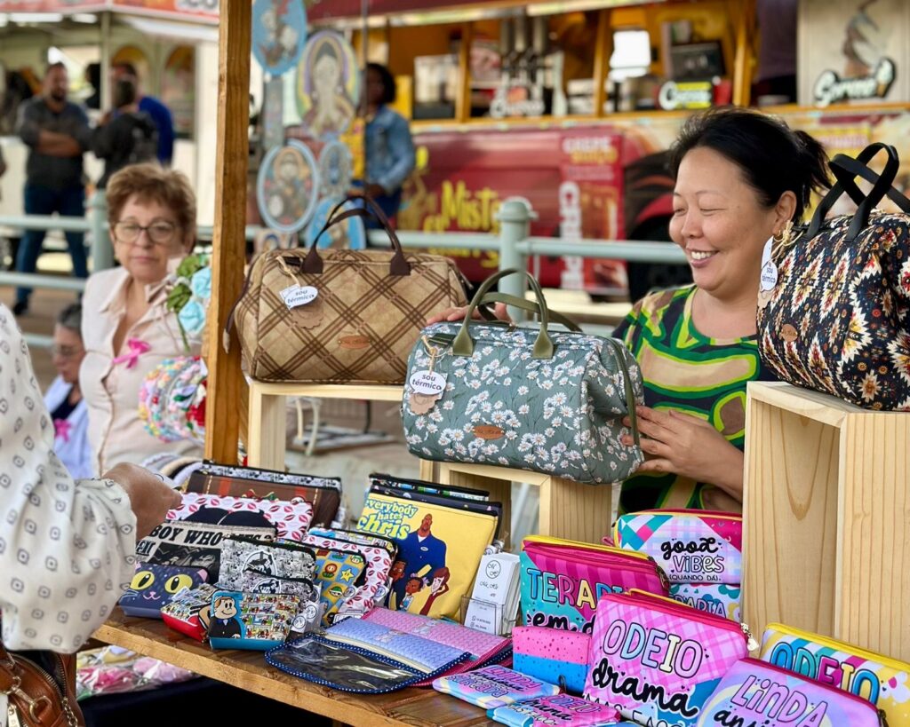 A imagem mostra uma feira com várias bolsas e nécessaires expostas em prateleiras de madeira. Uma mulher asiática sorri enquanto exibe uma bolsa com estampa geométrica. Ao seu lado, há uma bolsa verde com estampa de flores brancas e outra marrom xadrez. Abaixo, uma variedade de nécessaires coloridas com estampas divertidas e frases em português, como "ODEIO DRAMA" e "GOOD VIBES". Uma mulher mais velha, de óculos, está sentada em uma das barracas ao fundo. O ambiente parece ser ao ar livre, com outras pessoas e uma barraca de comida ao fundo.