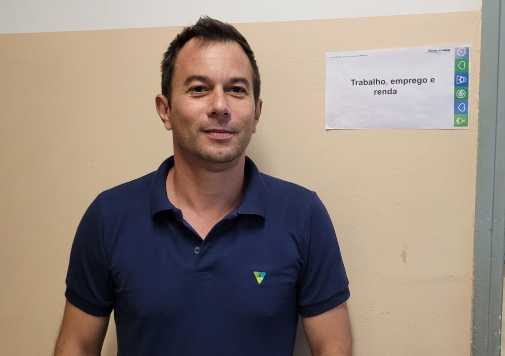 A imagem mostra um homem de pele clara, com cabelos castanhos curtos e um leve sorriso. Ele veste uma camiseta polo azul escura com um pequeno emblema triangular em verde e amarelo no lado esquerdo do peito. Ao fundo, há uma parede de cor bege e, à direita, um pedaço de papel colado na parede com o título "Trabalho, emprego e renda". O papel também contém um logotipo e alguns ícones verdes.