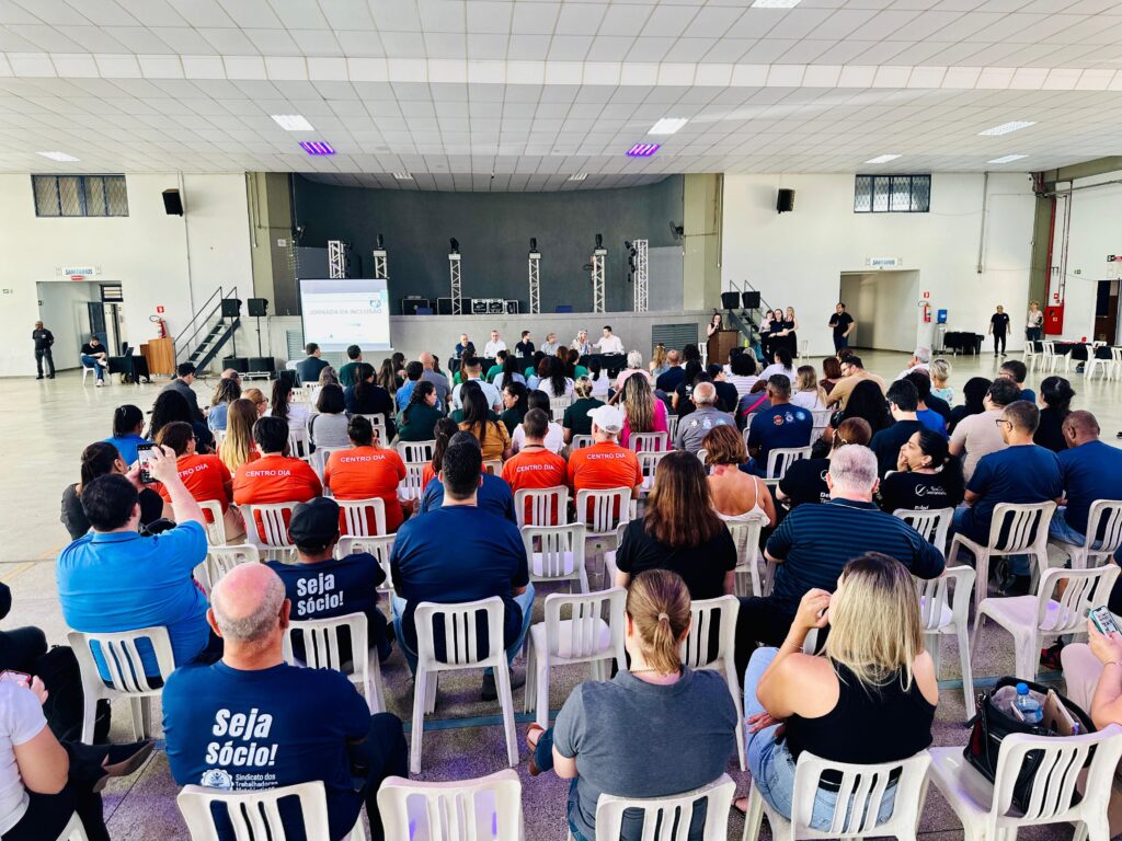 A imagem mostra um evento sendo realizado em um grande salão, possivelmente um auditório ou ginásio. Há um palco ao fundo com um telão exibindo a mensagem "JORNADA DA INCLUSÃO". Várias pessoas estão sentadas em cadeiras brancas, dispostas em fileiras, assistindo a uma apresentação ou palestra. Na plateia, muitas pessoas usam camisetas com a inscrição "CENTRO DIA" em laranja, e algumas usam camisetas azuis com a frase "Seja Sócio!" e o logo de um sindicato. Há também pessoas vestindo roupas casuais. No palco, um grupo de pessoas está sentado em uma mesa, provavelmente os palestrantes ou painelistas. Há também pessoas em pé, algumas apresentando e outras em pé na lateral. A iluminação do salão é feita por luzes fluorescentes no teto, com algumas luzes roxas decorativas em destaque. Nas paredes laterais, há sinalização para "SANITÁRIOS". A atmosfera geral sugere um evento comunitário ou corporativo focado em inclusão.
