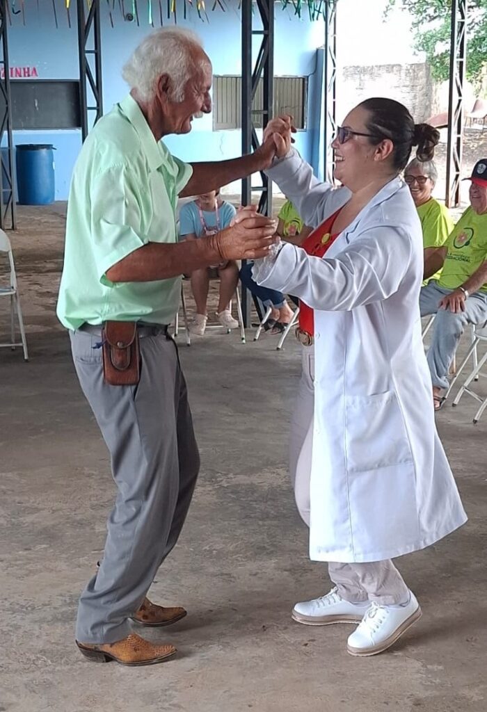 A imagem mostra um casal dançando, com o homem mais velho à esquerda e a mulher à direita. O homem, com cabelos brancos e vestindo uma camisa polo verde clara, está sorrindo e parece estar em movimento, com um pé ligeiramente levantado. Ele usa calças cinzas e botas de couro marrom com um padrão de escamas. Um pequeno coldre de couro está preso ao seu cinto. A mulher, com óculos escuros e um sorriso radiante, veste um jaleco branco de laboratório sobre uma roupa vermelha. Ela tem o cabelo preso em um coque e usa tênis brancos. Ela está segurando a mão do homem, e ambos parecem estar se divertindo muito enquanto dançam. Ao fundo, algumas pessoas estão sentadas em cadeiras, observando a dança. O ambiente parece ser um espaço aberto ou um salão com piso de concreto e algumas estruturas de metal e tecido decorativo no teto. Há também um barril azul no canto esquerdo da imagem.