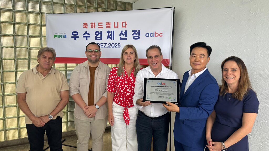 A imagem mostra seis pessoas em frente a um banner. O banner tem texto em coreano e português, com o logotipo da "PREFEITURA PIRA" e "acibc".As pessoas estão dispostas em duas fileiras. Na fileira de trás, da esquerda para a direita, estão um homem mais velho com cabelo claro e um homem mais jovem com óculos. Na fileira da frente, da esquerda para a direita, estão uma mulher com uma blusa vermelha com bolinhas brancas, um homem segurando uma placa de premiação, um homem em um terno azul e uma mulher com um vestido azul escuro.O homem que segura a placa de premiação parece ser o homenageado, pois o texto na placa diz "Liderança e Desenvolvimento" e "Helio Zanatta". A placa também tem o logotipo da "acibc".O fundo é uma parede com um padrão de blocos de vidro e uma parede texturizada. A iluminação parece ser interna, criando um ambiente formal para a ocasião. A atmosfera geral é de celebração e reconhecimento.