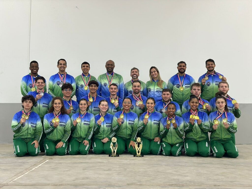 A imagem mostra um grupo de 23 pessoas, a maioria jovens, que parecem ser uma equipe desportiva, posando para uma foto em grupo. Todos estão vestidos com uniformes desportivos azuis e verdes, e a maioria exibe medalhas de ouro ao pescoço, sugerindo que venceram uma competição. Há dois troféus dourados em primeiro plano, um à esquerda e outro à direita, com a palavra "CAMPEÃO" escrita na base do troféu da esquerda, reforçando a ideia de que a equipe é campeã. A iluminação é uniforme, e o fundo é uma parede branca lisa, mantendo o foco nas pessoas e nas suas conquistas. A disposição em duas filas, com alguns membros ajoelhados à frente, é típica de fotos de equipe
